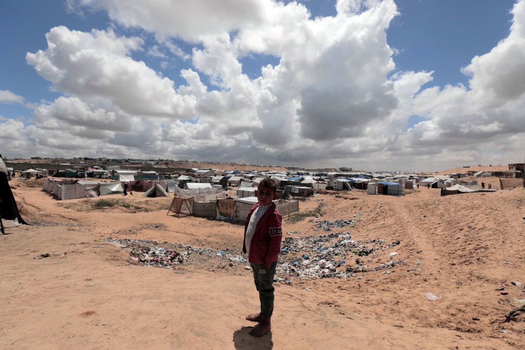 Au premier plan, un garçon pieds nus vêtu d'une veste rouge se tient devant un vaste campement de tentes de fortune étalé sur un sol sablonneux sous un ciel bleu nuageux.