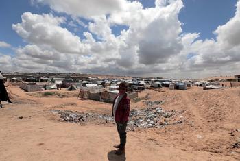 A barefoot boy in a red jacket stands in the foreground before a sprawling camp of makeshift tents spread across sandy ground under a cloudy blue sky.