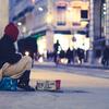 Une personne portant un bonnet rouge est assise sur un trottoir en ville la nuit, à côté d'une pancarte en carton et d'une tasse rouge.