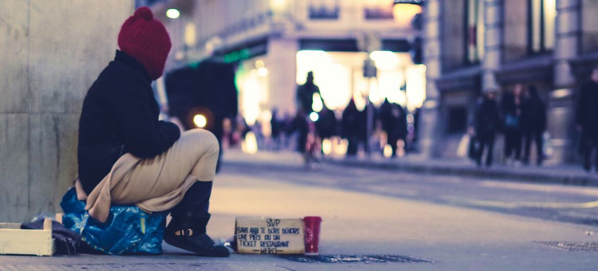 Une personne portant un bonnet rouge est assise sur un trottoir en ville la nuit, à côté d'une pancarte en carton et d'une tasse rouge.