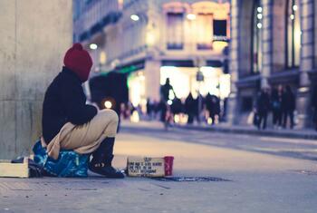 Une personne portant un bonnet rouge est assise sur un trottoir en ville la nuit, à côté d'une pancarte en carton et d'une tasse rouge.