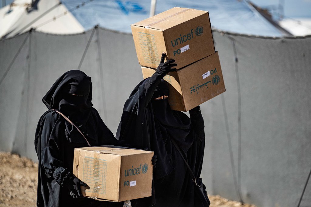 امرأتان ترتديان عباءات ونقابات سوداء تحملان صناديق مساعدات UNICEF في مخيم بسوريا.