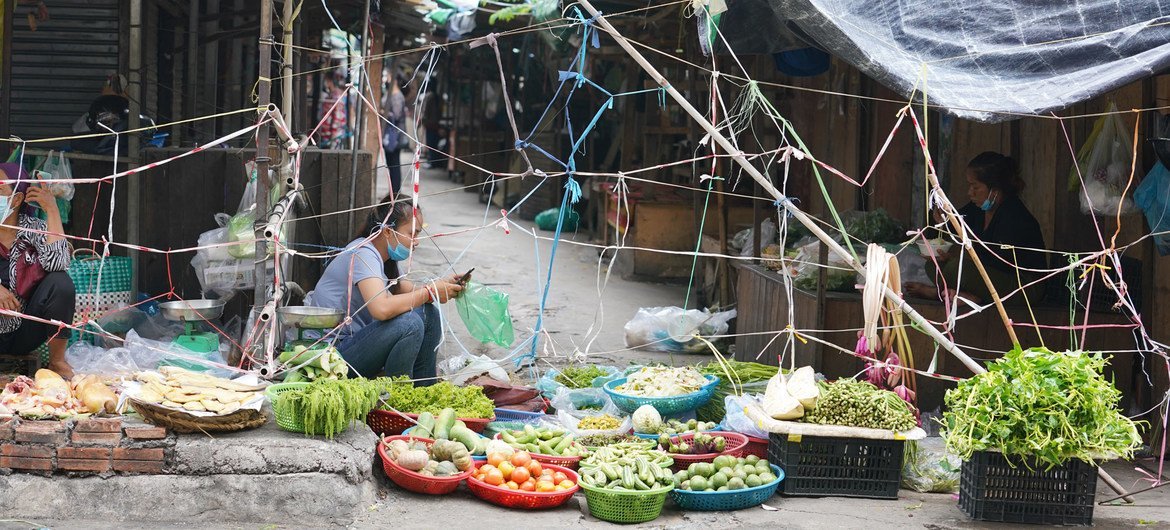 A woman wearing a face mask crouches beside a street-side display of fresh vegetables and fruits in colorful baskets at a makeshift local market stall covered by tarpaulin and bamboo poles.