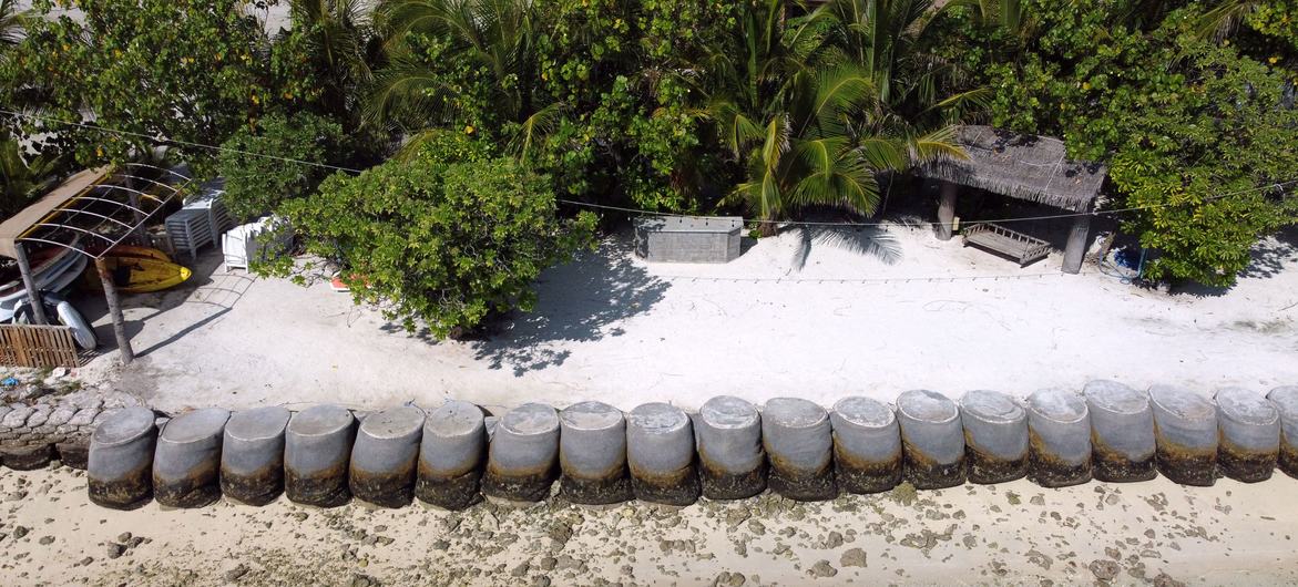Aerial view of a row of grey concrete cylinders lining the edge of a white sand beach on Dhiffushi island in the Maldives, with palm trees and a thatched-roof hut behind them.