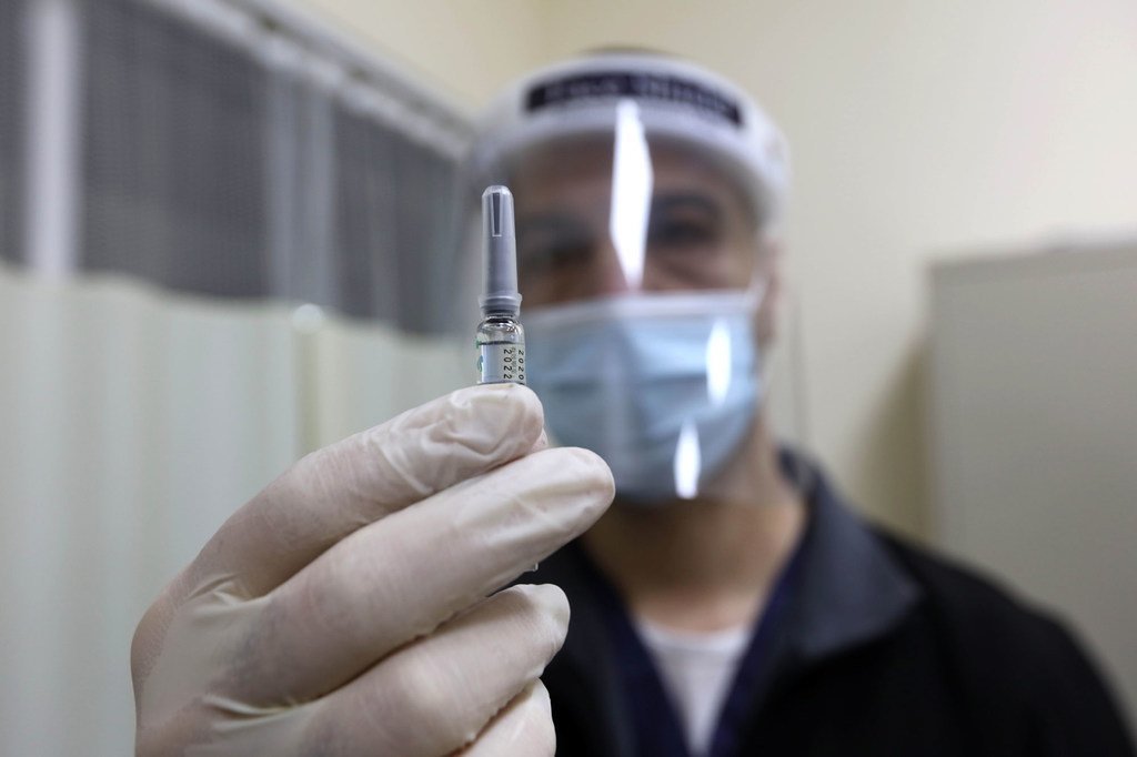 Un professionnel de santé en tenue de protection individuelle tient un flacon de vaccin au premier plan, son visage étant flou en arrière-plan.
