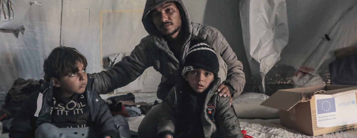 Un homme et deux jeunes garçons, vêtus de vêtements d'hiver, se trouvent à l'intérieur d'une tente dans un camp de réfugiés. Une boîte d'aide de UNICEF est visible dans un coin.