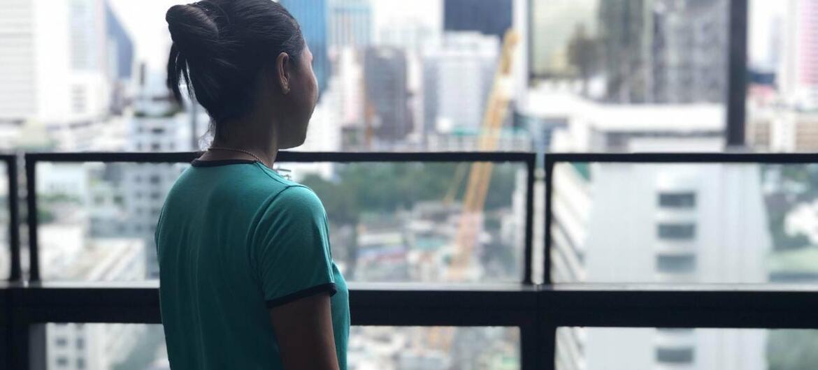 A woman migrant worker in Thailand stands by a window overlooking a cityscape, reflecting on her journey and experiences.