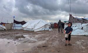 A young boy stands in a muddy displaced persons camp in Jabalya, North Gaza, holding a bucket. Tents are visible in the background under a cloudy sky.