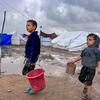 Dos niños desplazados caminan por un campamento fangoso con tiendas en el fondo, llevando baldes durante un descanso en la lluvia en Jabalya, en el norte de Gaza.