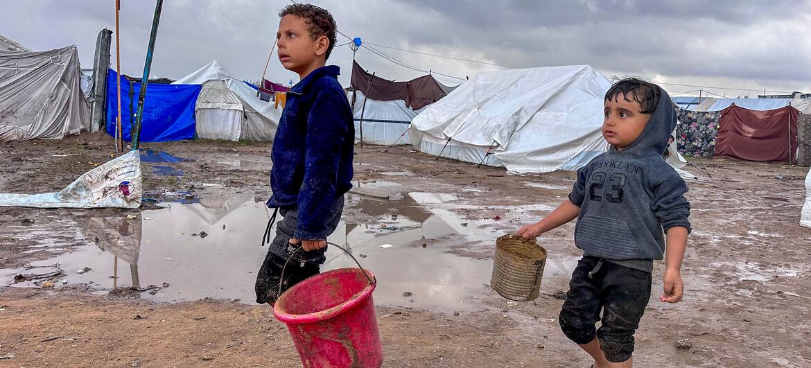 Dos niños desplazados caminan por un campamento fangoso con tiendas en el fondo, llevando baldes durante un descanso en la lluvia en Jabalya, en el norte de Gaza.