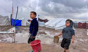 Dos niños desplazados caminan por un campamento fangoso con tiendas en el fondo, llevando baldes durante un descanso en la lluvia en Jabalya, en el norte de Gaza.