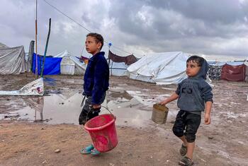 Dos niños desplazados caminan por un campamento fangoso con tiendas en el fondo, llevando baldes durante un descanso en la lluvia en Jabalya, en el norte de Gaza.