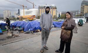 A man and woman stand in front of tents set up in a city centre, with construction cranes and buildings in the background. This depicts displaced people living in difficult conditions in Lebanon.