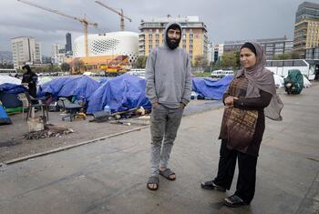 A man and woman stand in front of tents set up in a city centre, with construction cranes and buildings in the background. This depicts displaced people living in difficult conditions in Lebanon.