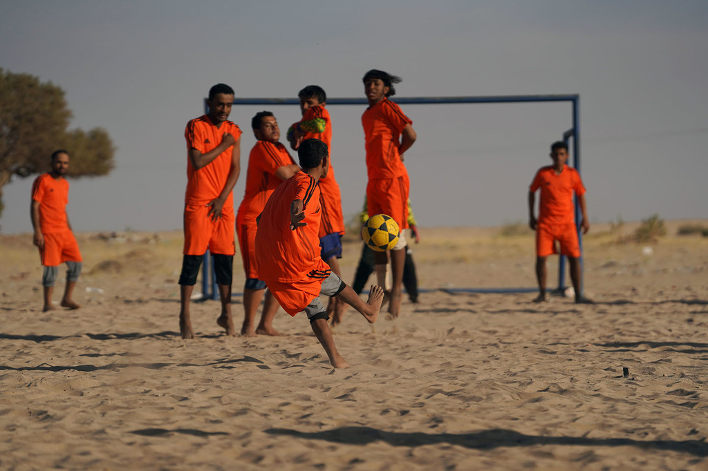 Un grupo de jóvenes con camisetas naranjas juegan al fútbol en un campo de arena en un entorno desértico.