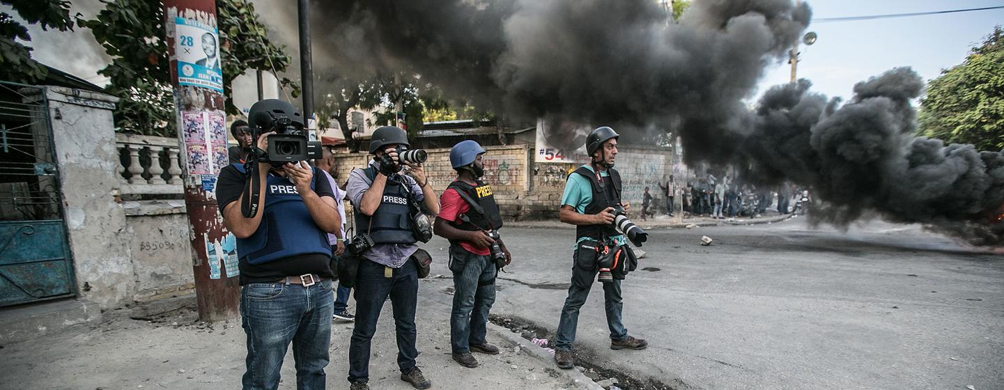 Fotoperiodistas con cascos y chalecos de prensa documentan una protesta en Puerto Príncipe, Haití, mientras el humo negro se eleva en el fondo.