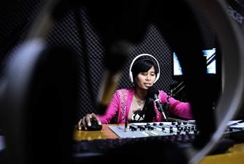 A female journalist wearing headphones speaks into a microphone at an audio mixing console in a radio studio, photographed by Chris Peken in Myanmar.