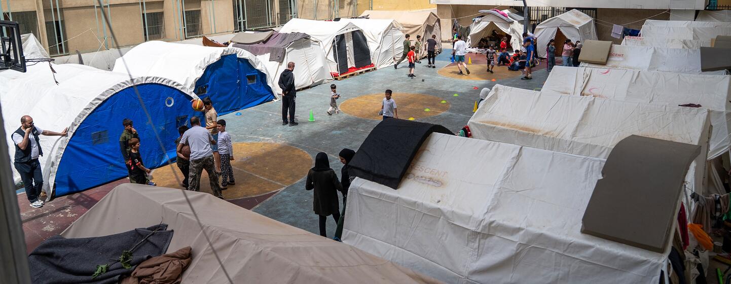Des enfants jouant au basket-ball et à d'autres jeux dans une cour entourée de tentes blanches et bleues, dans un abri collectif temporaire à Beyrouth, au Liban.