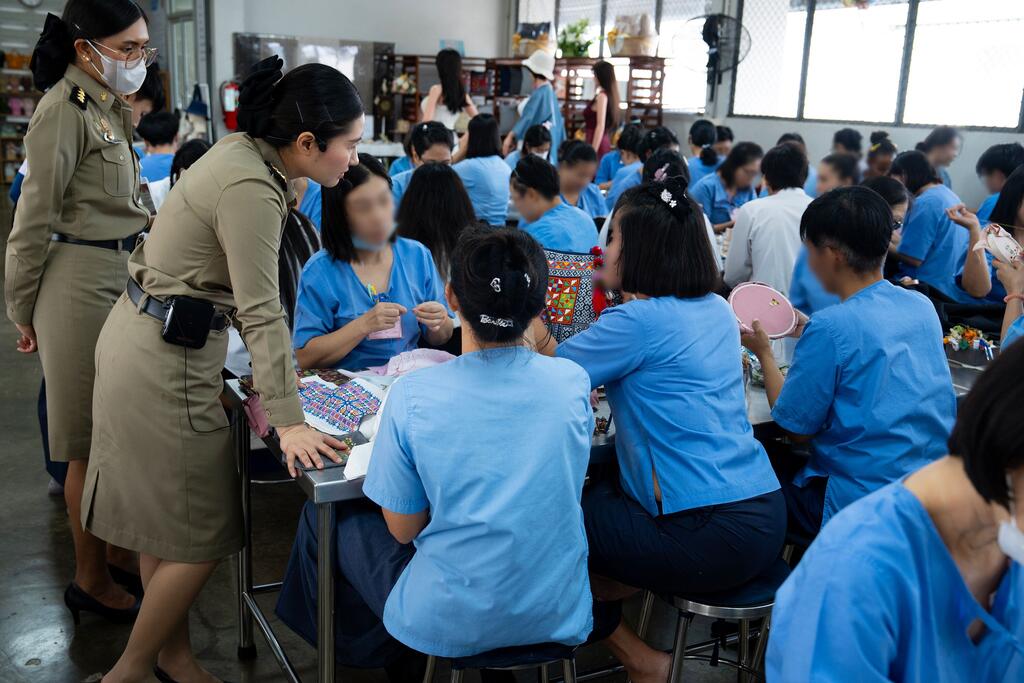 Female prison officers supervise female inmates in blue uniforms working on handicrafts in a prison workshop in Thailand.
