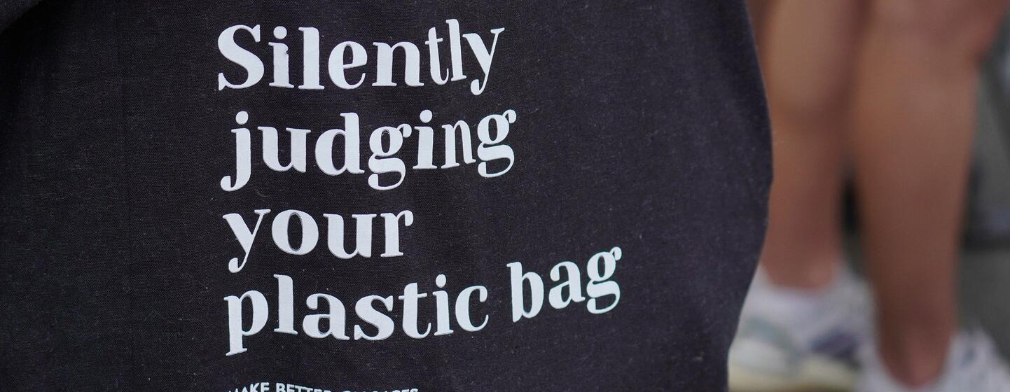 Primer plano de una bolsa negra con texto blanco que dice "Juzgando en silencio tu bolsa de plástico" y "TOMAR MEJORES DECISIONES".
