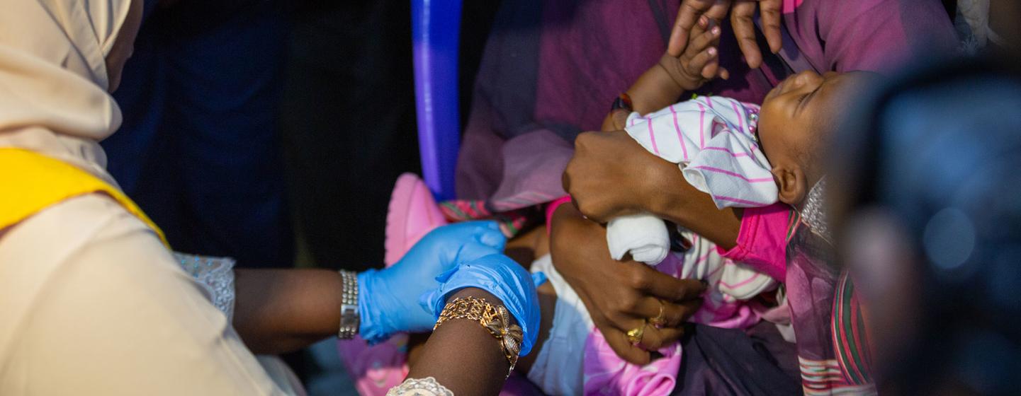 Un professionnel de la santé portant des gants bleus administre un vaccin à un bébé tenu par sa mère.