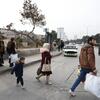 Civilian families, including children, walk through a damaged urban street in Aleppo, Syria, carrying bags as they evacuate during conflict.