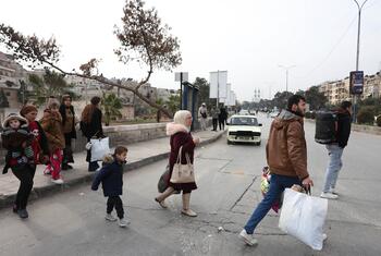 Civilian families, including children, walk through a damaged urban street in Aleppo, Syria, carrying bags as they evacuate during conflict.