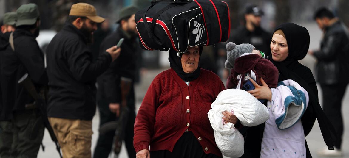 A woman carrying a large bag on her head and another woman holding a child walk through a street in Aleppo, Syria, as displaced civilians evacuate due to escalating hostilities.