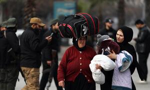 A woman carrying a large bag on her head and another woman holding a child walk through a street in Aleppo, Syria, as displaced civilians evacuate due to escalating hostilities.
