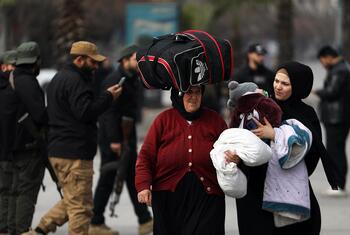 امرأة مسنة تحمل حقيبة سفر على رأسها وامرأة أخرى تحمل طفلا يرتدي ملابس شتوية، وعدد من الأشخاص في أحد شوارع حلب، في سوريا، أثناء مغادرتهم بسبب تصاعد الأعمال العدائية.