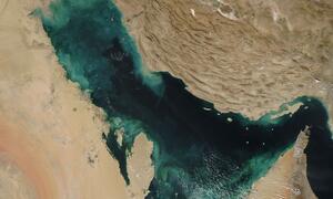 A satellite image from NASA's Aqua satellite shows the Persian Gulf, with tan and beige desert landmasses on either side and swirling patterns of green, blue, and tan in the water.