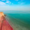 Una vista de la Playa Roja en la isla de Ormuz en Irán, con la distintiva costa de arena rosa y acantilados rojos que se encuentran con aguas turquesas bajo un cielo azul.