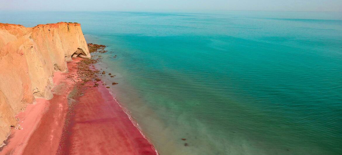 Uma vista da Praia Vermelha na ilha de Ormuz, no Irã, com a distintiva costa de areia rosa e penhascos vermelhos encontrando águas turquesa sob um céu azul.