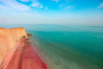Una vista de la Playa Roja en la isla de Ormuz en Irán, con la distintiva costa de arena rosa y acantilados rojos que se encuentran con aguas turquesas bajo un cielo azul.