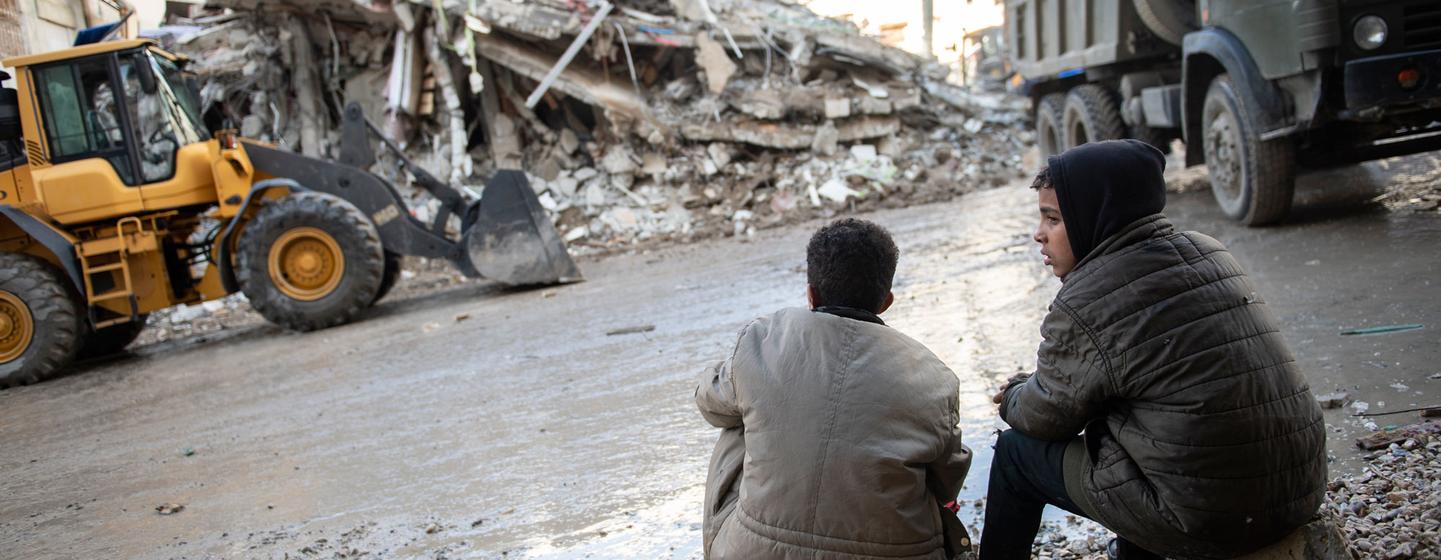 Deux jeunes garçons sont assis sur des décombres dans une rue de Jableh, en Syrie, regardant une excavatrice dégager les débris d'un bâtiment effondré après le tremblement de terre de 2023.