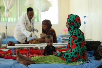 A woman in a green and red patterned headscarf sits on a hospital bed with a young child in Dollow, Somalia, while medical staff treat other patients in the background.