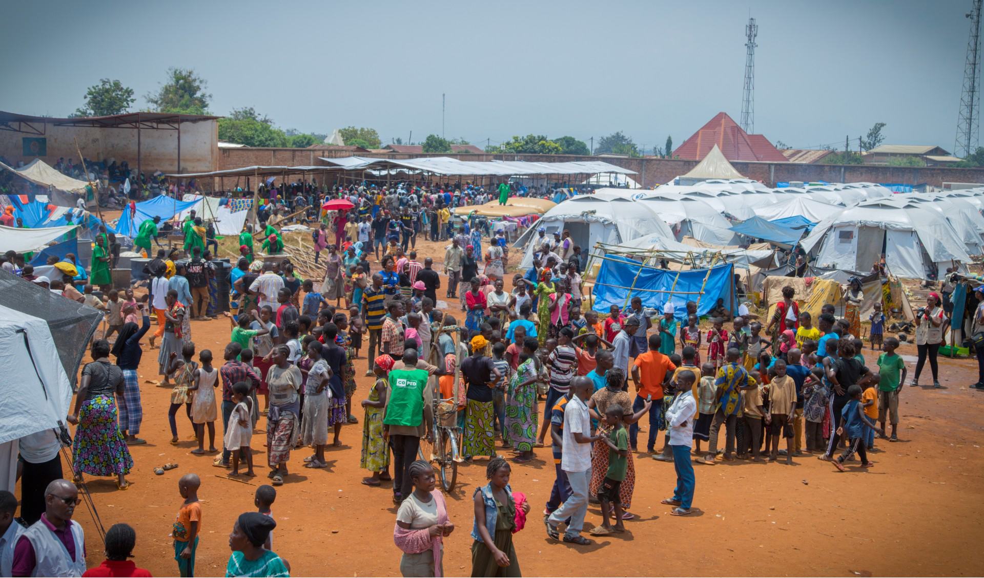 Les familles et les enfants déplacés se rassemblent dans un camp temporaire de réfugiés au Burundi, après avoir fui le conflit en République démocratique du Congo. Le camp comporte des rangées de tentes blanches et bleues sous un ciel clair, avec des gens qui marchent et interagissent dans une zone poussiéreuse et ouverte.