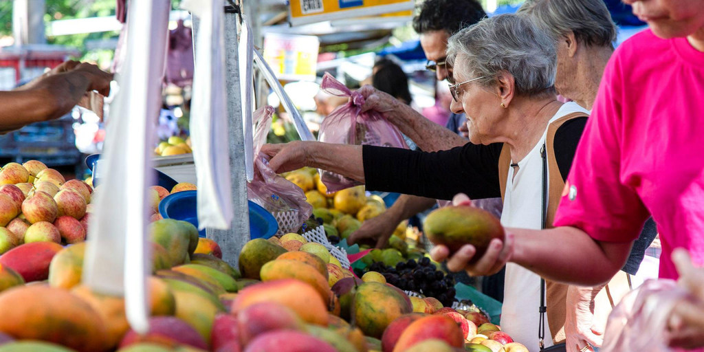 Una anciana con el pelo gris y gafas elige un mango en un bullicioso mercado de frutas y verduras al aire libre.