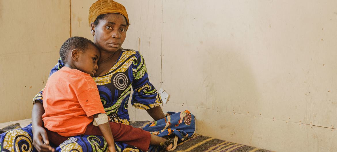 Dans une chambre simple, une femme et un jeune enfant sont assis sur un lit. La femme, vêtue d'une robe à motifs et d'un foulard, regarde l'objectif en tenant l'enfant contre elle.
