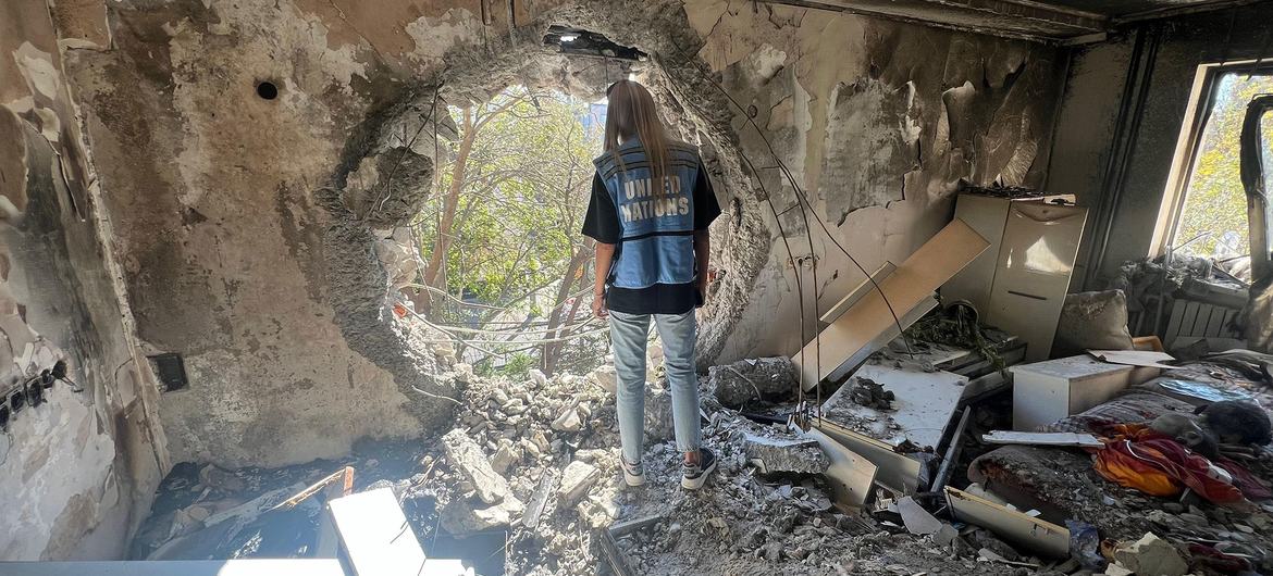 A UNOCHA aid worker stands in a destroyed building in Ukraine, looking through a large hole in the wall at trees outside.