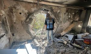 A UNOCHA aid worker stands in a destroyed building in Ukraine, looking through a large hole in the wall at trees outside.