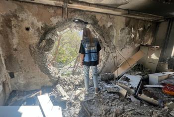 A UNOCHA aid worker stands in a destroyed building in Ukraine, looking through a large hole in the wall at trees outside.