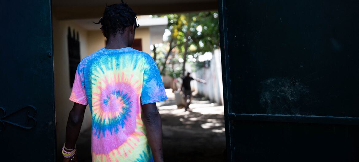 A young person wearing a colorful tie-dye shirt walks through an open doorway in Haiti, with another figure visible in the background, reflecting efforts to prevent child recruitment and support reintegration.