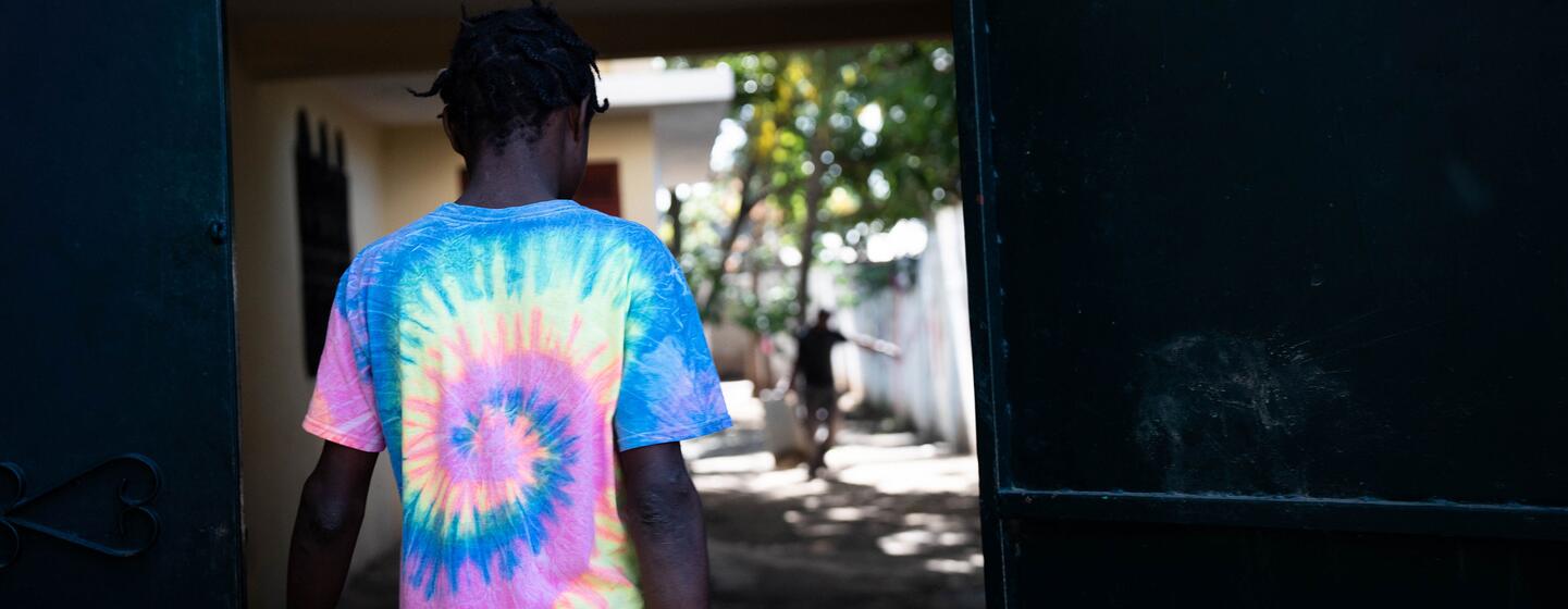 Um jovem vestindo uma camisa colorida de gravata passa por uma porta aberta no Haiti, com outra figura visível no fundo, refletindo os esforços para evitar o recrutamento de crianças e apoiar a reintegração.