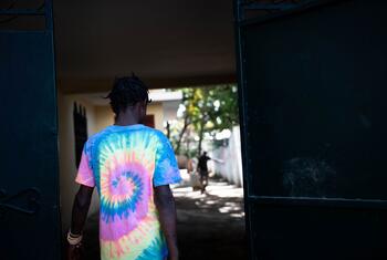 Um jovem vestindo uma camisa colorida de gravata passa por uma porta aberta no Haiti, com outra figura visível no fundo, refletindo os esforços para evitar o recrutamento de crianças e apoiar a reintegração.