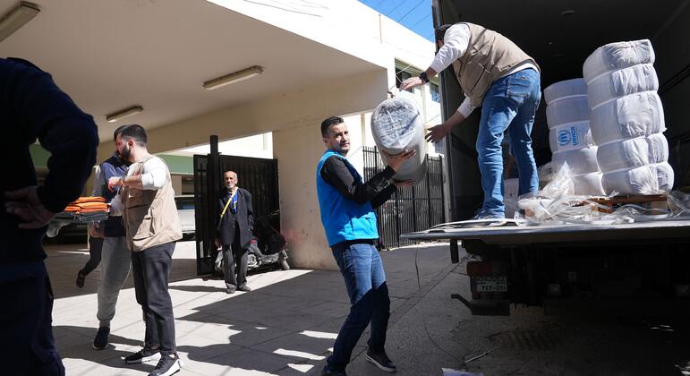 ممثل المفوضية السامية للأمم المتحدة لشؤون اللاجئين وموظفو الوكالة الوطنية لشؤون اللاجئين يوزعون مواد إغاثة أساسية للعائلات النازحة في ملجأ جماعي في أدرا، محافظة سعيدة، لبنان. رجال يرتدون سترات يطردون إمدادات إغاثة من شاحنة.