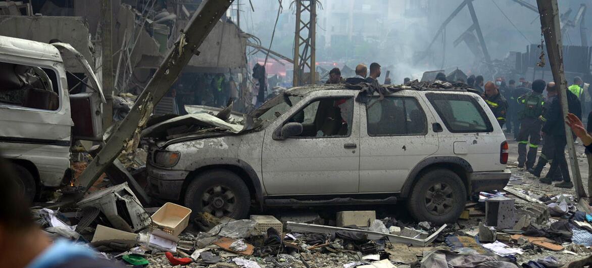 A scene of destruction in Beirut following airstrikes, with a damaged white SUV in the foreground amidst rubble and debris, while emergency responders work in the background.