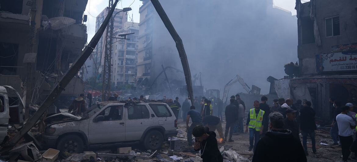 A scene of devastation in Beirut following airstrikes, showing collapsed buildings, smoke rising from rubble, and emergency personnel assessing the damage.