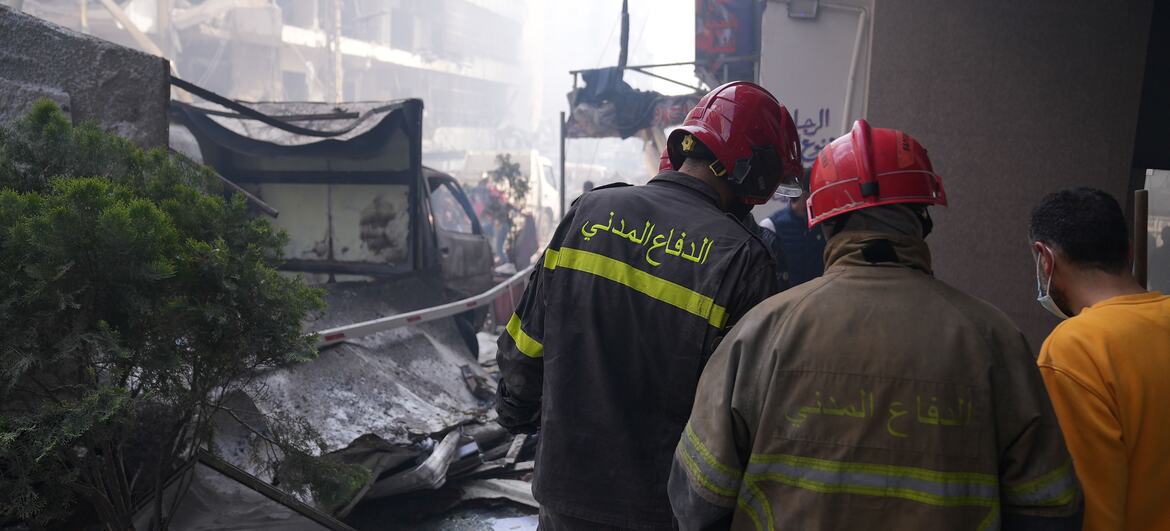 Two firefighters in red helmets and uniforms with Arabic script on the back survey a scene of destruction with rubble and damaged buildings in Beirut, Lebanon.