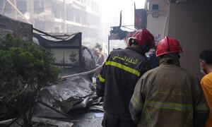 Two firefighters in red helmets and uniforms with Arabic script on the back survey a scene of destruction with rubble and damaged buildings in Beirut, Lebanon.